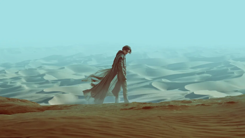 Paul Atreides, avanzando con el mítico destiltraje de la saga, un mono sellado herméticamente y diseñado para favorece el reciclaje del agua del propio cuerpo. Avanza de lado, el desierto de Arrakis está de fondo